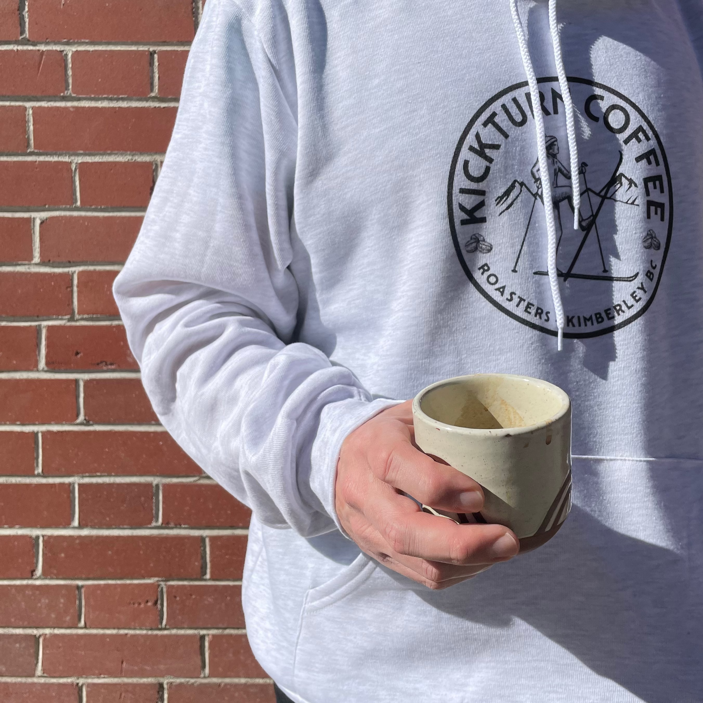 Person wearing a hoodie with Kickturn Coffee logo holding a ceramic cup against a brick wall.